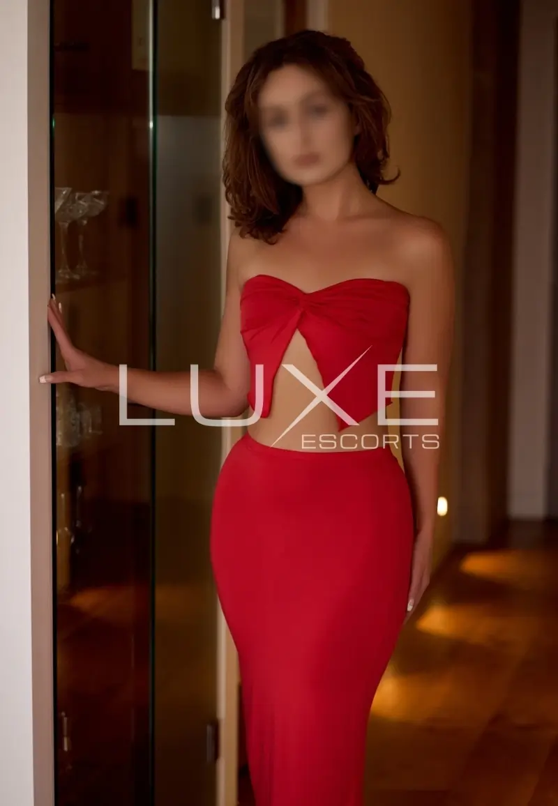 Elegant Brisbane escort in a red evening dress standing confidently in a softly lit luxury apartment